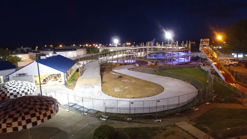 Floodlit night aerial of a curving concrete go-kart track, a lit violet spiral and a blue-roofed pit building